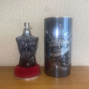 Jean Paul Gaultier Le Male Tattoo EdT - Cool parfymflaska i form av en manlig torso i mörkgrått glas med tatueringsinspirerat motiv. Kommer med original metallburk och röd bas. Märket och designen ger en edgy och unik känsla. Perfekt för dig som gillar statement-dofter och snygg inredningsdetalj.