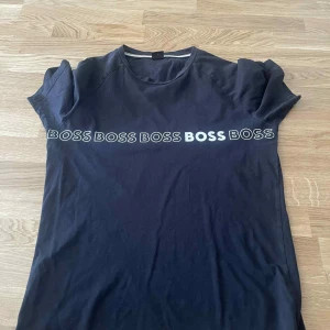 Svart BOSS t-shirt med logga - Svart t-shirt från BOSS i slim fit med klassisk rund hals och korta ärmar. Framsidan har flera BOSS-loggor i vitt tryck över bröstet. Tillverkad i mjuk bomull som känns skön mot huden. Perfekt för en clean och stilren look.