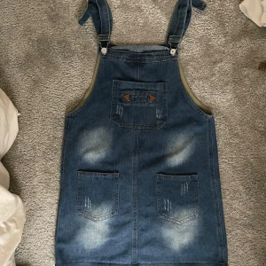 Jeansklänning med hängslen och fickor - Snygg jeansklänning i klassisk blå denim med justerbara hängslen och tre fickor framtill. Klänningen har slitna detaljer och dekorativa sömmar samt bruna läderdetaljer. Perfekt för en avslappnad och trendig look.