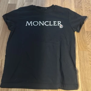 Svart Moncler t-shirt med logga - Svart t-shirt från Moncler med stor vit logotyp tryckt över bröstet och klassisk rund hals. T-shirten har korta ärmar och en enkel, stilren design som passar perfekt till jeans eller shorts.
