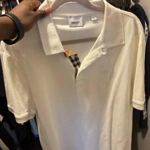 Vit pikétröja från Burberrry - Klassisk vit pikétröja från Burberryy med korta ärmar och ikonisk rutig detalj vid knäppningen. Tröjan har broderad B-logga på bröstet och är tillverkad i bomull för en skön och luftig känsla. Perfekt för en clean och stilren look.