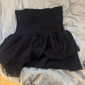 Svart volangkjol - Supersöt svart kortkjol med bred smock i midjan och dubbla volanger. Kjolen har en lätt och luftig känsla, perfekt för att styla med en crop top eller stickad tröja. Tunn, något transparent struktur i tyget ger extra detalj.