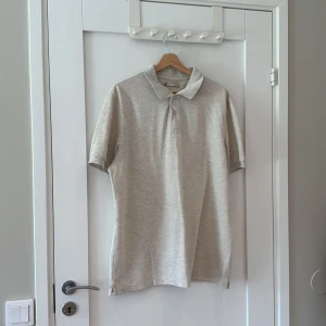 Beige pikétröja 2XL från Dressman - Klassisk beige pikétröja från Dressman i storlek 2XL. Tröjan har korta ärmar, krage och knäppning med två knappar framtill. Tillverkad i mjuk bomull som känns skön mot huden. Perfekt för en clean och avslappnad stil.