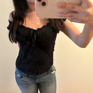 Brandy Melville Svart offshoulder blus med snörning  - Säljer en svart blus med offshoulder och korta ärmar. Toppen har en söt snörningsdetalj framtill och volangkant längs urringningen. Perfekt att styla med jeans eller kjol för en trendig look.