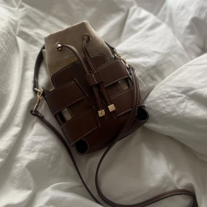 Brun och beige axelväska med snörning - Säljer en snygg axelväska i brun skinn med beige canvasdetaljer. Väskan har cool snörning upptill och justerbar axelrem. Perfekt storlek för mobilen, plånboken och lite till. Stilren design med kontrastsömmar och guldiga detaljer.