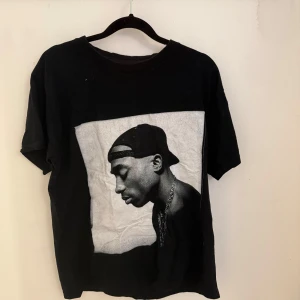 Svart t-shirt med tryck av artist - Svart t-shirt med stort svartvitt tryck framtill föreställande en känd artist i keps och kedja. Klassisk passform med rund hals och korta ärmar. Perfekt för dig som gillar streetstyle och vill sticka ut med en cool grafisk look.