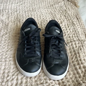 Svarta sneakers från Adidas, strl 39⅓ - Klassiska svarta sneakers från Adidas med vit sula och svarta snören. Skorna har en stilren design med de ikoniska tre ränderna på sidan och Adidas-logga på plösen. Tillverkade i skinnliknande syntetmaterial och passar perfekt till en streetwear-look.
