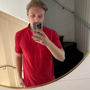 Röd pikétröja från Polo Ralph Lauren - Säljer en klassisk röd pikétröja från Polo Ralph Lauren i storlek M. Tröjan har korta ärmar, krage och knappar framtill samt den ikoniska blå loggan broderad på bröstet. Perfekt för en clean och sportig look.