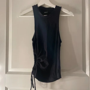 Svart linnetopp med snörning - Snygg svart ärmlös linnetopp från Bik Bok med rund halsringning och cool snörningsdetalj på sidan som ger en trendig look. Toppen är i mjuk bomull och har en tight passform som framhäver figuren. Perfekt att styla med jeans eller kjol för en modern vibe. Det är en storlek L men passar storlek M och kanske till och med storlek S. Den är också aldrig använd.