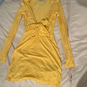 Gul omlottklänning med knyt från NA-KD - Snygg gul kort omlottklänning med lång ärm och knyt framtill. Klänningen har en v-ringad hals och är gjord i ett mjukt, lätt material som passar perfekt för varmare dagar.