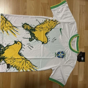 Vit Brasil Nike fotbollströja M - Snygg vit Brasilien fotbollströja från Nike med gröna och gula detaljer i krage och ärmslut. Stor gul papegojgrafik på framsidan, Brasilien-emblem och Nike-logga. Tillverkad i Dri-FIT material som andas och håller dig torr. Perfekt för dig som älskar fotboll och vill sticka ut. Ny med tags!