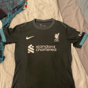 Liverpool Salah 11 Nike fotbollströja - Snygg svart Liverpool fotbollströja från Nike med Salah 11 på ryggen. Tröjan har korta ärmar med turkosa detaljer och klubbmärke samt sponsortryck på bröstet. Tillverkad i lätt och ventilerande funktionsmaterial, perfekt för match eller träning.