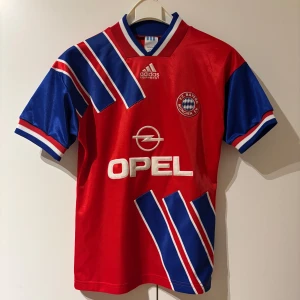 Bayern München retro fotbollströja Adidas - Klassisk Bayern München fotbollströja från Adidas med OPEL-logga på bröstet. Tröjan har korta ärmar, klubbmärke på vänster bröst och coola diagonala mönster i blått och vitt. Tillverkad i glansigt syntetmaterial. Tröjan är från säsongen 1993/94 och kostar runt 700kr på nätet. Hör av dig vid frågor! 