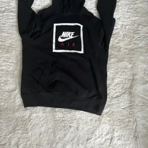 Svart Nike Air hoodie med logga - Svart hoodie från Nike Air med stor vit och röd logga på bröstet. Klassisk känguruficka framtill, huva och långa ärmar. Perfekt för dig som gillar streetwear och vill ha en stilren look med sportig vibe.