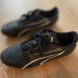 Svarta fotbollsskor från Puma strl 38 - Svarta fotbollsskor från Puma med vita detaljer och klassisk logga på plösen. Skorna har snörning och mönstrad sula med fasta dobbar för bra grepp på planen. Snygg, sportig design som sticker ut på matchen.