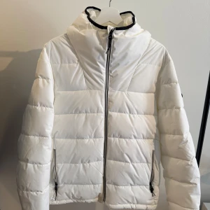 Vit vinterjacka med huva från Pull & Bear - Snygg vit vinterjacka med huva och svarta detaljer vid dragkedjan. Jackan har quiltad design, två fickor med dragkedja och en skön, vadderad känsla. Perfekt för kalla dagar när du vill hålla stilen enkel men ändå sticka ut. Märke: Pull & Bear