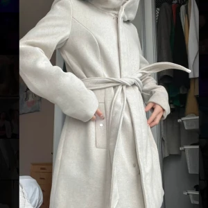 Beige kappa med huva och bälte - Snygg beige kappa med huva och knytskärp i midjan. Lång modell med dragkedja och knappar framtill, samt diskreta fickor på sidorna. Perfekt för kyligare dagar och ger en clean och stilren look. Köptes för 1200kr
