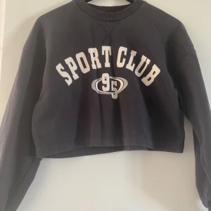 Svart croppad sweatshirt Gina Tricot - Svart croppad sweatshirt från Gina Tricot med trycket 'SPORT CLUB 95' framtill. Tröjan har långa ärmar, rund halsringning och är gjord i mjuk bomullsblandning. Perfekt för en sportig och chill look.