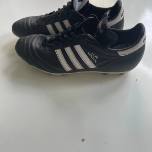 Adidas Copa Mundial fotbollsskor - Klassiska Adidas Copa Mundial fotbollsskor i svart skinn med vita detaljer. Skorna har snörning och rund tå, perfekt för dig som vill ha stilrena och tidlösa skor på planen. De är i storlek 46.