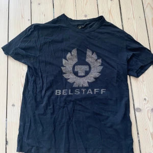 Svart Belstaff t-shirt med logga - Svart t-shirt från Belstaff med stor, grå logga framtill. Klassisk rund hals och korta ärmar. T-shirten är i mjuk bomull. Skick 8/10, storlek M men passar mer som  M/S