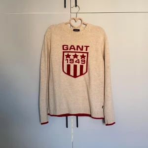 Beige stickad GANT tröja med rött tryck - Snygg beige stickad tröja från GANT med rött broderat logotyp-tryck och texten '1949' framtill. Tröjan ribbade muddar Perfekt för dig som gillar klassisk college-stil.