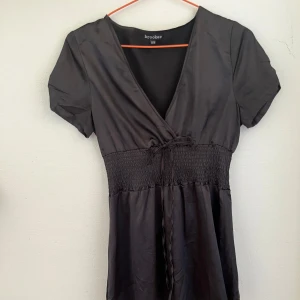 Svart miniklänning  - Svart kortklänning från Brooker med omlott-v-ringning, korta ärmar och smockad midja med knytband. Klänningen har en lätt glansig yta och faller snyggt. Perfekt för dig som gillar en enkel men stilren look.