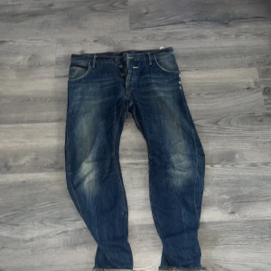 Blå jeans från G-Star RAW med dragkedjor - Säljer ett par blå jeans från G-Star RAW med coola detaljer som dragkedjor på bakfickorna och läderpatch i midjan. Jeansen har en snygg tvätt med slitningar och knäppning fram. Perfekta för dig som gillar streetstyle och vill sticka ut.