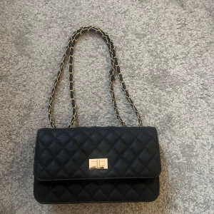 Svart quiltad axelväska med kedja - Stilren svart axelväska med quiltat mönster och guldfärgad kedja i axelremmen. Väskan har ett klassiskt lock med snyggt guldfärgat spänne framtill. Perfekt för dig som gillar tidlös design och vill ha en väska som sticker ut.