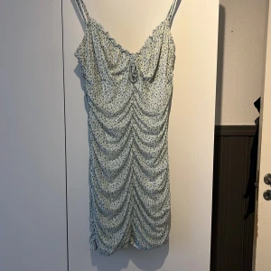 Blommig ljusblå/ljusgrön kortklänning med snörning - Supersöt kort klänning med ljusblå botten och små blommor i blått och grönt. Smala axelband, rynkade detaljer framtill och snörning vid bysten. Perfekt för varma dagar och har en riktigt fräsch sommarkänsla.