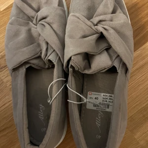 Beige slip-on sneakers med rosett - Snygga slip-on sneakers från Allegy i beige mocka-look med bred rosettdetalj framtill. Skorna har vit platt sula och rund tå, vilket ger en clean och trendig vibe. Perfekta för dig som gillar en stilren men ändå lekfull look.