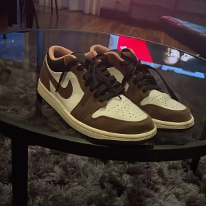 Nike Air Jordan 1 Low Mocha 40.5 - Nike Air Jordan 1 Low i snygg mocha-färg med bruna och vita paneler, svart swoosh och svarta skosnören. Klassisk Air Jordan-logga på hälen och mjuk beige insida. Perfekt för dig som vill sticka ut med en ikonisk sneaker.