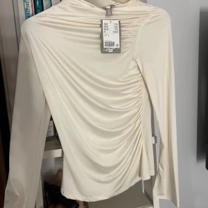Krämvit topp - H&M - Superfin krämvit tilo från H&M med lång ärm och snygg. Toppen har en lätt glansig finish och är tillverkad i ett stretchigt syntetmaterial. Perfekt för dig som gillar trendiga detaljer och stilren look.