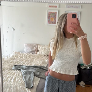 Vit smockad topp med volangkant - Supersöt vit topp med smockad midja och volangkant nertill. T-shirtrmodell med korta ärmar och rund halsringning. Perfekt för dig som gillar en feminin och trendig stil. Mjuk och skön bomullskänsla.