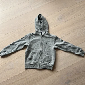 Grå hoodie från Polo Ralph Lauren - Snygg grå hoodie från Polo Ralph Lauren med dragkedja och klassisk broderad logga på bröstet. Tröjan har huva, känguruficka och ribbade muddar. Perfekt för chill dagar och enkel att matcha med jeans eller joggers.