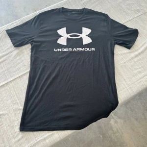 Svart Under Armour t-shirt loose fit - Svart t-shirt från Under Armour med stor vit logga och text på bröstet. Klassisk rund halsringning och korta ärmar. Modellen är loose fit och tillverkad i mjuk bomull, perfekt för en sportig och avslappnad stil.
