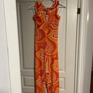 Orange swirl JUMPSUIT från Amisu - Snygg långklänning från Amisu med färgstarkt swirlmönster i orange, rosa, gult och vitt. Klänningen har halterneck, öppen rygg och smock i midjan för en bekväm passform. Perfekt för sommarens alla tillfällen och ger riktiga retrovibbar!