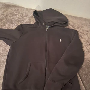 Svart hoodie från Polo Ralph Lauren - Svart hoodie från Polo Ralph Lauren med dragkedja och klassisk logga broderad på bröstet. Tröjan har huva, långa ärmar och två fickor framtill. Perfekt för en chill och stilren look. Materialet är mjuk bomullsmix som känns skön mot huden. Frakt sker samma dag om inte dagen efter!👍Vid snabb affär pris går att diskuteras!
