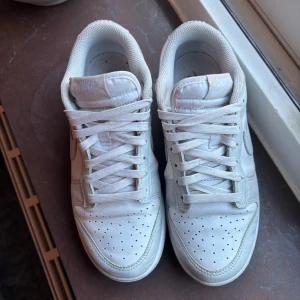 Nike Dunk Low sneakers - Snygga Nike Dunk Low sneakers i helvitt skinn med klassisk siluett och perforerad tå. Skorna har vit sula, snörning och diskret Nike-logga på hälen och sidan. Perfekta för dig som gillar en clean och stilren look.