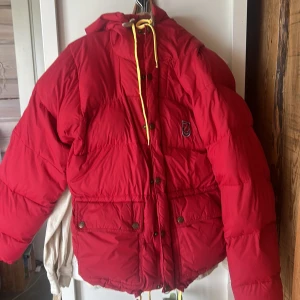 Röd dunjacka från Fjällräven xxs (herr) - Röd Fjällräven med huva och gula snören.  stora fickor och huva. Storleken är en herr xs och sitter som en dam. Dock finns en fläck , se sista bilden.