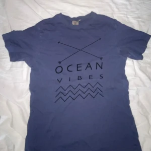 Blå t-shirt Tribute Ocean Vibes S - Blå t-shirt från Tribute i storlek S med svart tryck 'Ocean Vibes', korsade pilar och vågmönster framtill. Klassisk rund hals och korta ärmar. Skön och mjuk bomullskvalitet, perfekt för en chill och avslappnad stil.