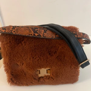 Brun fluffig axelväska River Island - Säljer en brun axelväska från River Island med mjuk pälsimitation och detaljer i svart skinn. Väskan har ett brett axelband med ormskinnsmönster och guldfärgat spänne. Insidan har flera fack och dragkedja. Perfekt för dig som vill sticka ut med en unik accessoar.