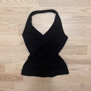 Svart halterneck-topp  - Snygg svart linne med v-ringad hals och smala axelband. Toppen är figurnära och har en enkel, stilren design som passar perfekt till jeans eller kjol. Liten spetskant längs halsringningen för en extra touch.