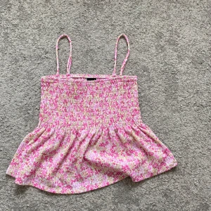 Blommig linnetopp från Gina Tricot - Supersöt blommig linnetopp från Gina Tricot i rosa, vit och lite grönt. Smockad överdel med tunna axelband och volangig nederdel. Perfekt för varma dagar när du vill ha en luftig och trendig look.