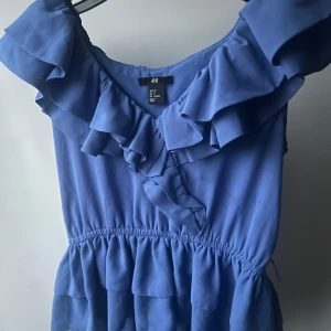 Blå volang topp från H&M - Supersöt blå topp från H&M med volanger längs urringningen och midjan. Toppen har en luftig känsla och resår i midjan för en snygg passform. 