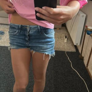 Blå jeansshorts från ONLY - Snygga blå jeansshorts från ONLY med låg midja och råa, fransiga kanter. Klassisk femficksmodell med dragkedja och knapp framtill. Perfekta för sommardagar och har en cool, avslappnad vibe. Materialet är jeans med lätt tvättad effekt.