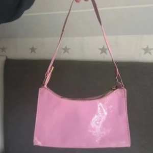 Glansig rosa axelväska - Säljer en söt axelväska i glansigt rosa material med enkel design och guldfärgade metalldetaljer. Väskan har en smidig dragkedja upptill och ett kort axelband som gör den perfekt att slänga över axeln. Perfekt för att addera lite färg till din outfit!
