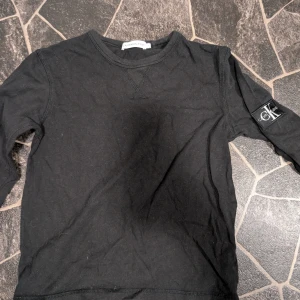 Svart långärmad tröja Calvin Klein Jeans stl 10år - Svart långärmad tröja från Calvin Klein Jeans med rund halsringning och snyggt CK-märke på ärmen. Tröjan är i mjuk bomull och har en clean, stilren look som passar perfekt till jeans eller kjol.