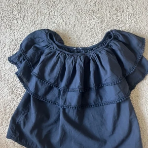 Mörkblå offshoulder volangblus - Supersöt mörkblå blus med offshoulder-modell och dubbla volanger runt hela överdelen. Blusen har en luftig passform och är dekorerad med små bollar längs volangkanterna. Perfekt för dig som gillar en romantisk och trendig stil. Har storlek m/l men väldigt liten i modellen skulle säga att den passar mer S/M