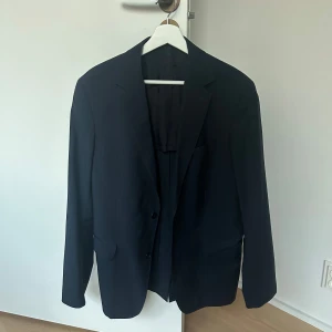 Mörkblå randig kavaj med knappar - Snygg mörkblå kavaj med diskreta ränder och klassisk passform. Kavajen har två knappar framtill, tre knappar vid ärmslut och fickor med lock. Perfekt för dig som vill ha en stilren och tidlös look.