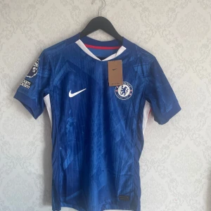 Chelsea 25/26 hemmatröja Nike Delap #9 tagg sitter på! - Chelsea FC hemmatröja från Nike med Delap och nummer 9 på ryggen. Tröjan är blå med vita och röda detaljer, korta ärmar och klubbmärke på bröstet. Tillverkad i dri-fit material, perfekt för match eller träning. 